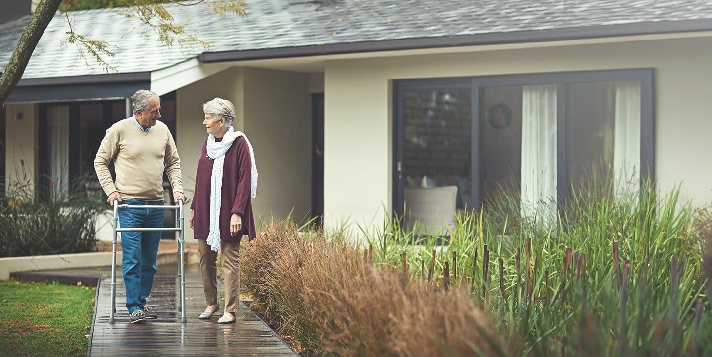 Ein älterer Mann mit Gehhilfe zusammen mit einer Frau vor einem Haus