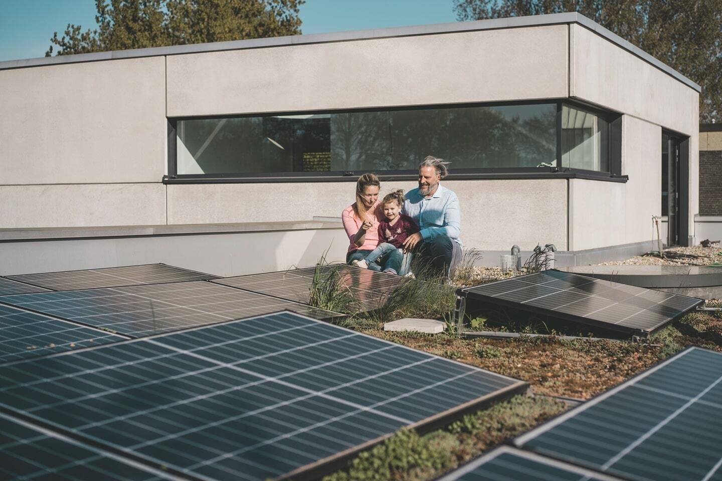 Mutter, Vater und Kind sitzen auf dem Dach eines modernen Wohnhauses und betrachten eine Solaranlage.
