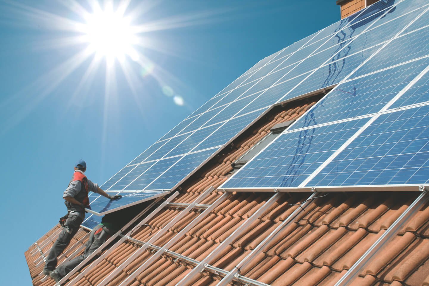 Bauarbeiter stehen vor strahlend blauem Himmel und Sonnenstrahlen auf einem Dach und montieren eine Solaranlage