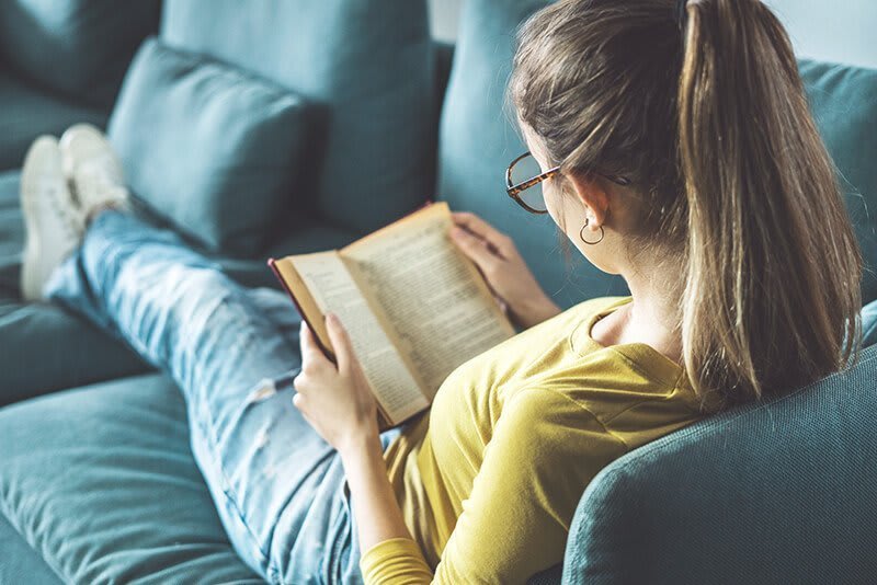 Das Bild zeigt eine Frau von hinten, die auf einem blauen Sofa sitzt und ein Buch liest. 