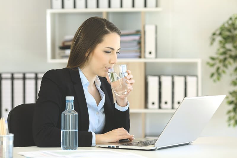 Frau sitzt am Schreibtisch mit Laptop, Wasserflasche und trinkt einen Schluck aus Glas.